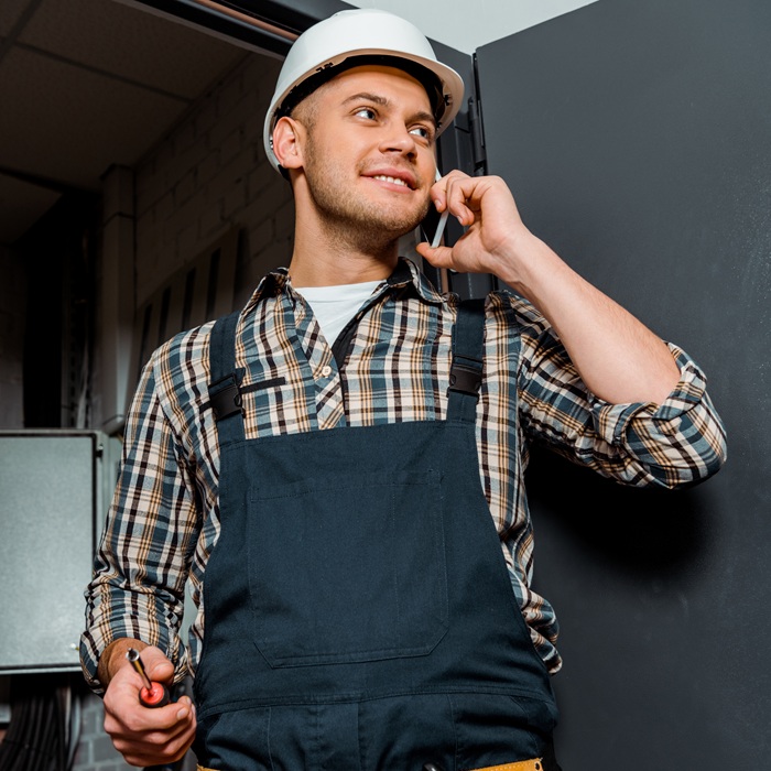 cheerful installer in safety helmet holding screwdriver while talking on smartphone