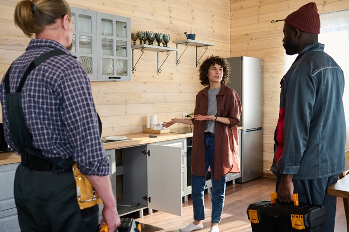 Young brunette woman in casualwear describing technical problem with kitchen pipe to intercultural plumbers from repair service company