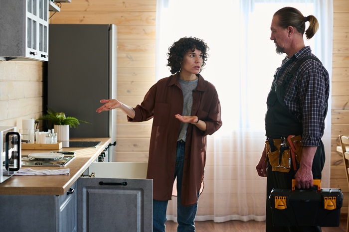 Young worried housewife looking at plumber and explaining him problem with pipe or faucet while standing by kitchen counter with open cabinet