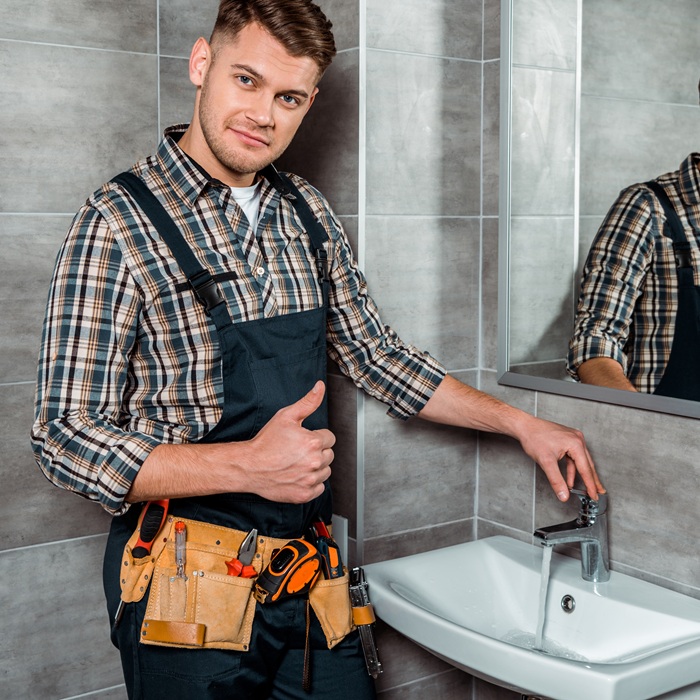 handsome installer standing near faucet with flowing water and showing thumb up