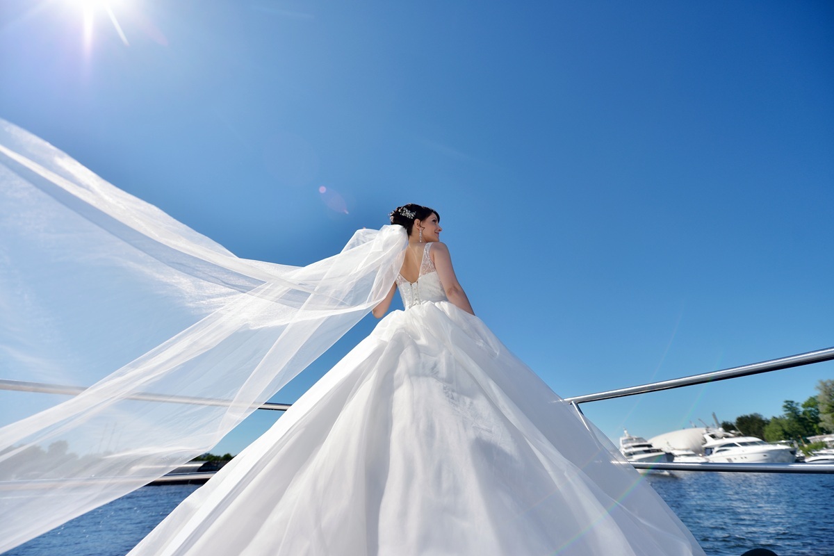 Beauty bride in bridal gown with lace veil on a yacht. Beautiful model girl in a white wedding dress. Female portrait on the nature. Woman with hairstyle. Cute lady outdoors